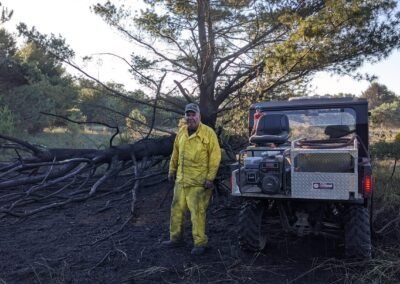 A member in yellow wildland gear stands beside the department’s UTV and its pump unit on burned ground