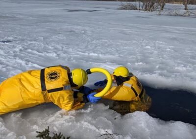 A rescuer in an ice rescue suit reaches a person in the water with a rescue pole at a hole in the ice